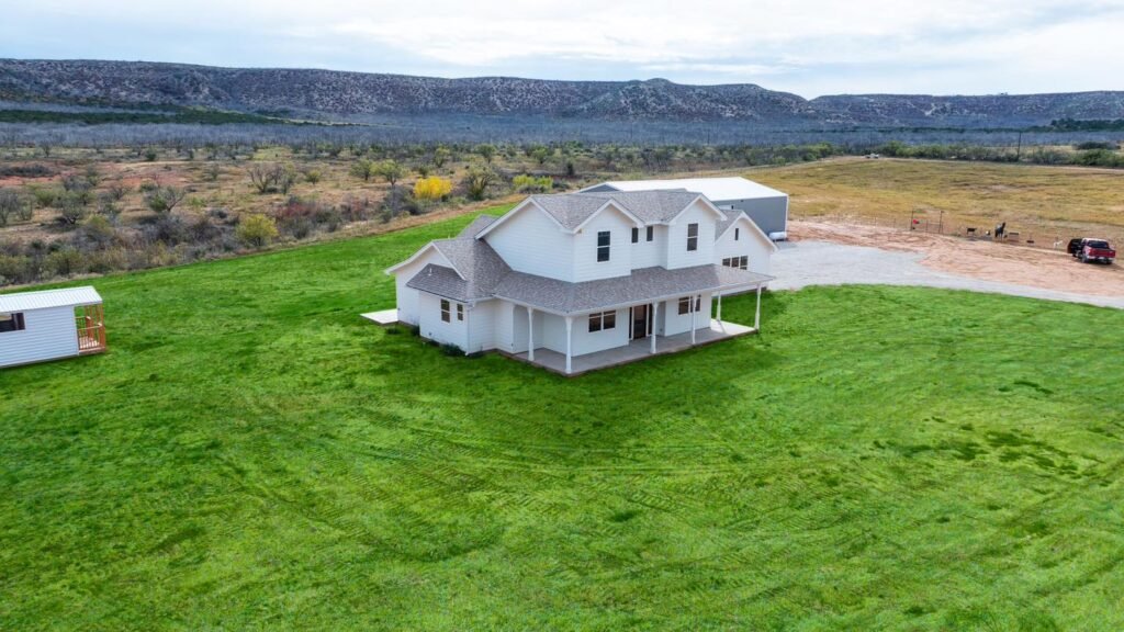 Drone view of a white custom home in Abilene, TX on a large green property with a detached shop building and West Texas hills in the background.