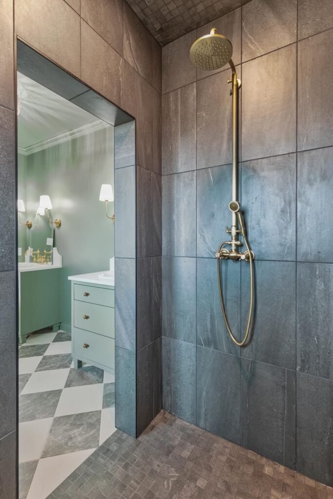 Modern walk-in shower with large gray tile walls and brass rain showerhead, opening to bathroom with green vanity and patterned floor tile.